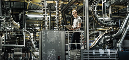 Fabrikinnere mit Förderbändern und einem Mitarbeiter auf einer Plattform, der einen Laptop benutzt