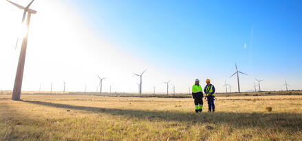 Zwei Mobil-Ingenieure stehen mitten in einem Windpark. Mehrere Windkraftanlagen in einer Reihe
