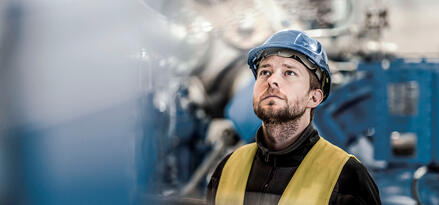Mann mit Schutzhelm und gelber Sicherheitsweste, der in einer Industrieanlage nach oben schaut